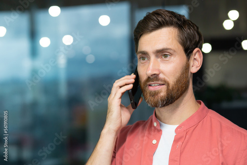 Successful businessman in a red shirt talking on the phone inside the office, a man with a beard at the workplace is talking to a client, a satisfied boss in casual clothes.