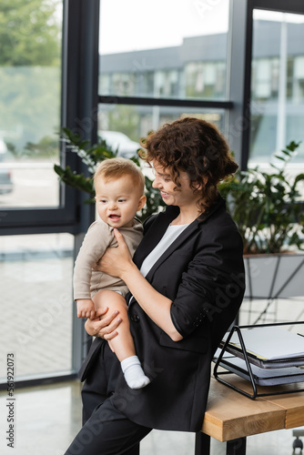 businesswoman in black suit holding little child near work desk with documents in office.