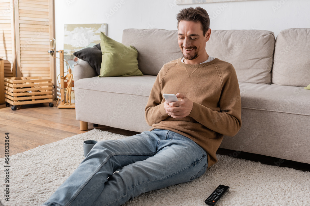 happy man in beige jumper messaging on smartphone while sitting on carpet at home.