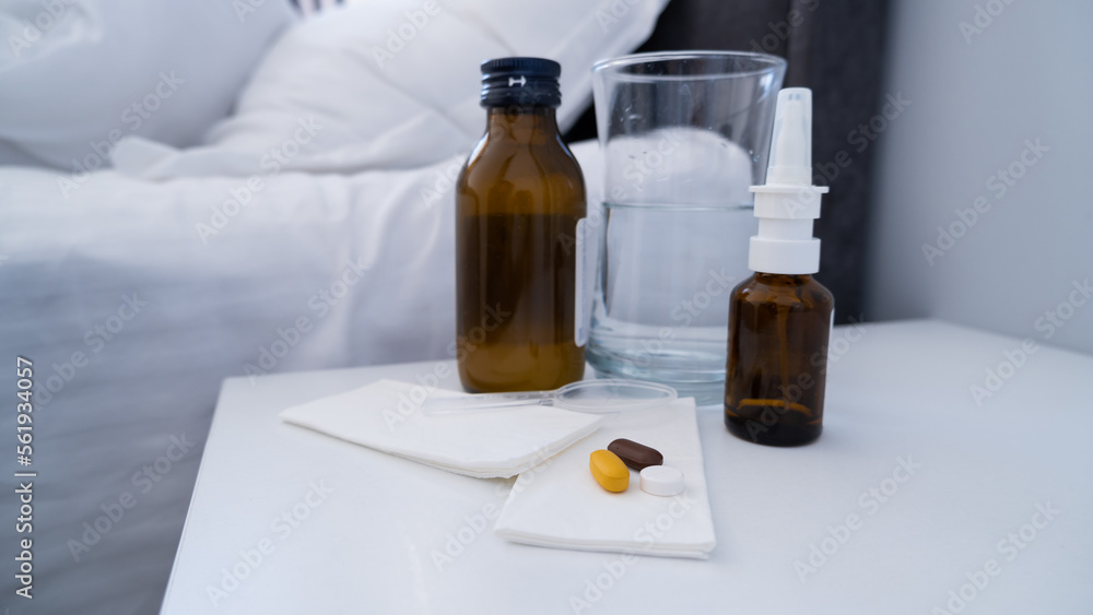 Medicine, pills, bottle of syrup, nasal spray and a glass of water on the white bedside table. Illness treatment Medicine, pills, bottle of syrup, nasal spray and a glass of water on the white bedside table. Illness treatment