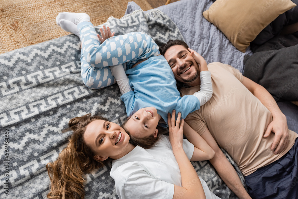 top view of positive parents and cheerful kid lying on bed at home.