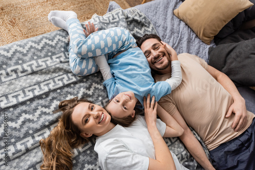 top view of positive parents and cheerful kid lying on bed at home.
