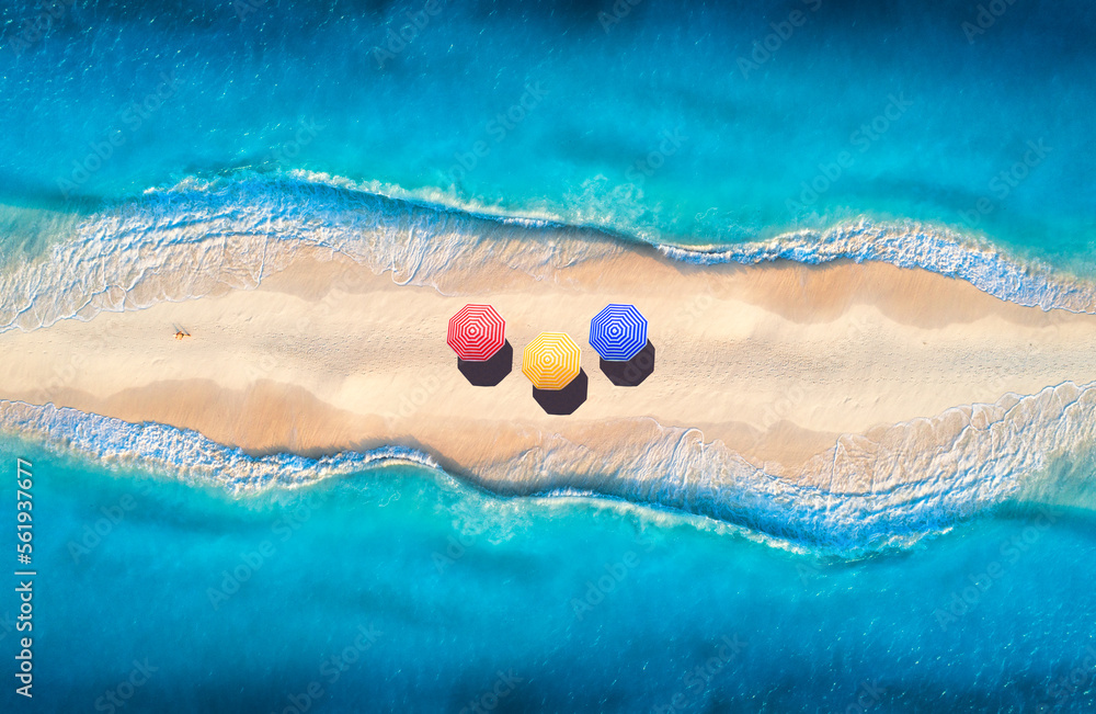 Obraz premium Aerial view of transparent blue sea with waves on the both sides and sandy beach with colorful umbrellas at sunset. Top view of sandbank. Summer travel. Tropical landscape with white sand and ocean