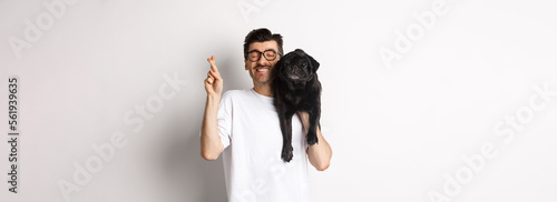 Hopeful smiling dog owner making a wish, holding cute black pug on shoulder and cross fingers for good luck, white background photo