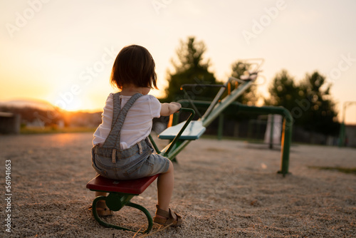 back view of one small caucasian toddler child sitting alone on the seesaw in park in sunset lonely with no friends copy space childhood growing up concept social issues rejected photo
