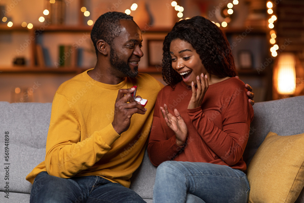 Man surprising his girlfriend with an engagement ring on the couch at home in the living room Man surprising his girlfriend with an engagement ring on the couch at home in the living room