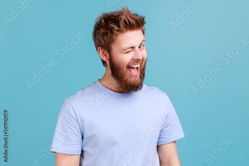 Portrait of bearded man being in good mood, smiling broadly and winking at camera, keeps mouth open, having flirting facial expression. Indoor studio shot isolated on blue background. photo