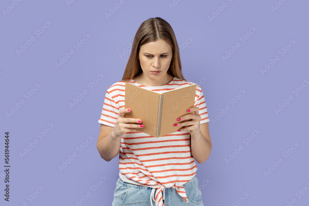 Portrait of serious concentrated blond woman wearing striped T-shirt holding and reading book, being impressed by plot, reads with attention. Indoor studio shot isolated on purple background. Portrait of serious concentrated blond woman wearing striped T-shirt holding and reading book, being impressed by plot, reads with attention. Indoor studio shot isolated on purple background.