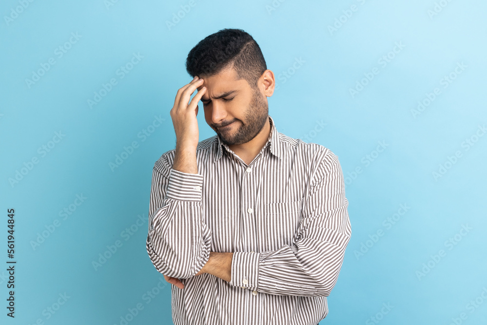 Businessman with beard making facepalm gesture keeping hand on head, blaming himself for bad memory, unforgivable mistake, wearing striped shirt. Indoor studio shot isolated on blue background. Businessman with beard making facepalm gesture keeping hand on head, blaming himself for bad memory, unforgivable mistake, wearing striped shirt. Indoor studio shot isolated on blue background.
