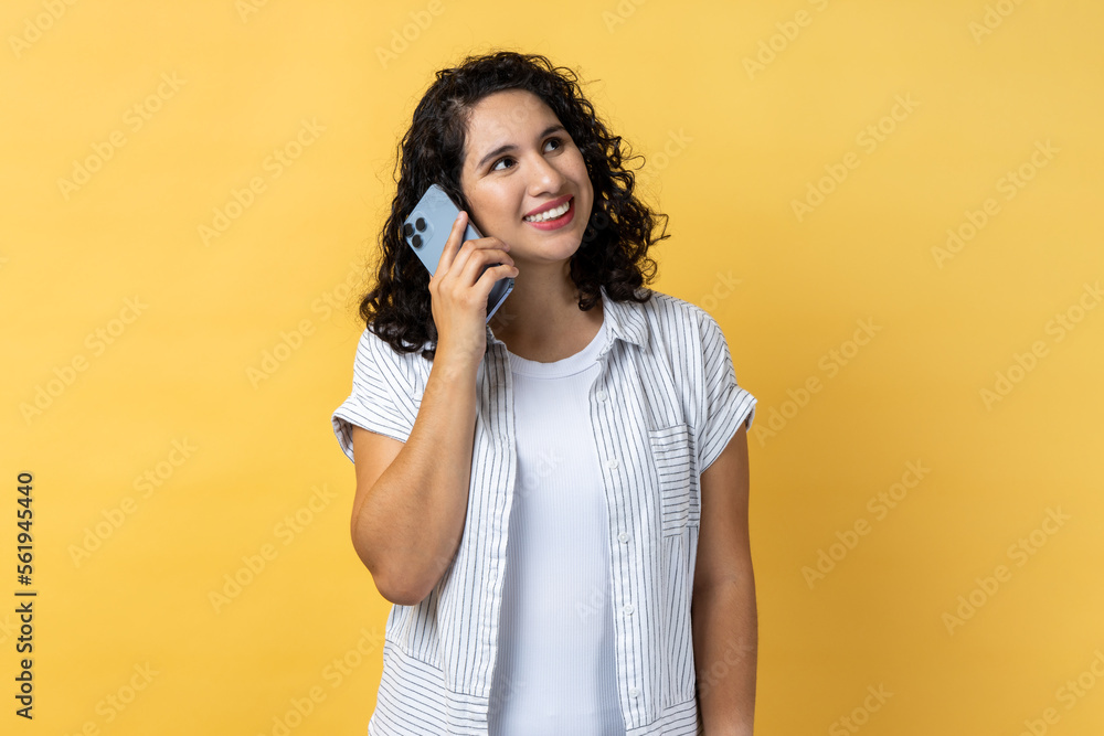 Portrait of smiling satisfied young adult beautiful woman with dark wavy hair standing and talking on smartphone, hearing good news. Indoor studio shot isolated on yellow background. Portrait of smiling satisfied young adult beautiful woman with dark wavy hair standing and talking on smartphone, hearing good news. Indoor studio shot isolated on yellow background.