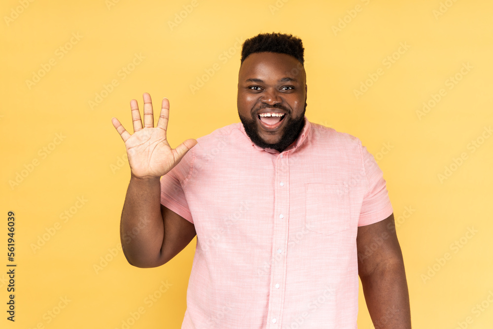 Portrait of friendly bearded man in pink shirt greeting you rising hand and waving, saying hi, glad to see you, looking at camera with toothy smile. Indoor studio shot isolated on yellow background. Portrait of friendly bearded man in pink shirt greeting you rising hand and waving, saying hi, glad to see you, looking at camera with toothy smile. Indoor studio shot isolated on yellow background.