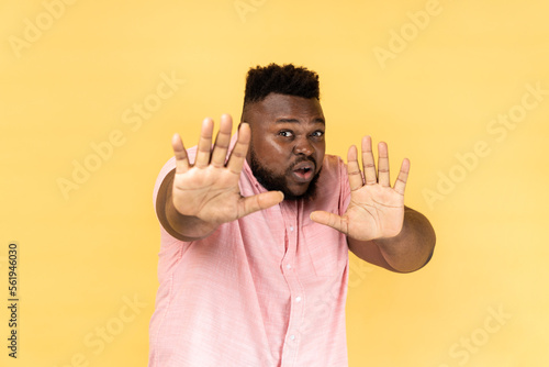 No, it's scary. Portrait of frightened shocked man wearing pink shirt raising hands in fear, looking horrified and panicking, hiding from phobia. Indoor studio shot isolated on yellow background.