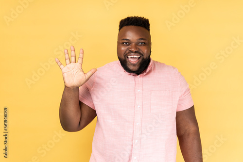 Portrait of friendly bearded man in pink shirt greeting you rising hand and waving, saying hi, glad to see you, looking at camera with toothy smile. Indoor studio shot isolated on yellow background.