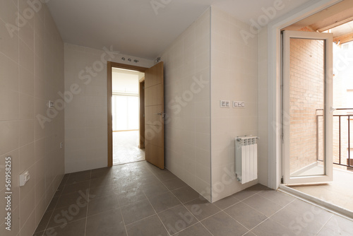 Empty room to install a kitchen with a clothesline terrace with a white aluminum door and partition, an oak access door, a white aluminum radiator and dark stoneware floors © Toyakisfoto.photos