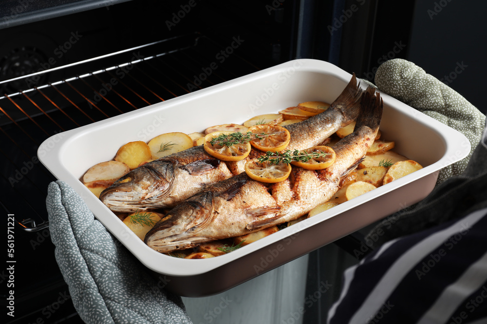 Woman taking out baking tray with sea bass fish and potatoes from oven - obrazy, fototapety, plakaty 
