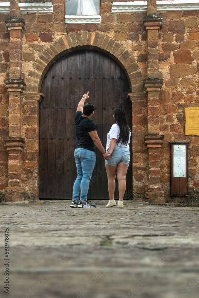Pareja de jovenes novios tomados de la mano frente a la capilla del pueblo. Hombre señalando hacia arriba - Charalá, Santader, Colombia