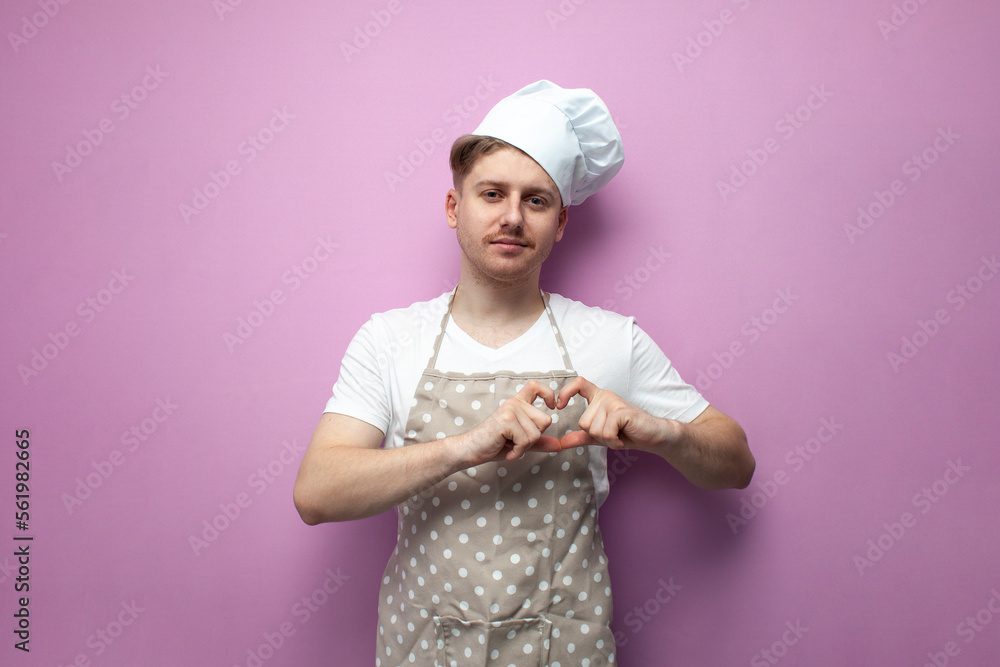 guy housewife in an apron and a hat poses and shows a heart sign with his fingers on a pink background, love for cooking