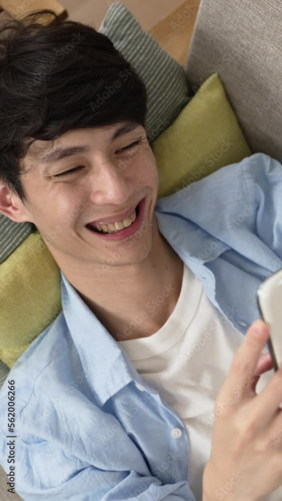 Vertical Screen: view from above of a cheerful asian male lying on the sofa and smiling with happiness while sending text message to his friend on the phone at home Vertical Screen: view from above of a cheerful asian male lying on the sofa and smiling with happiness while sending text message to his friend on the phone at home