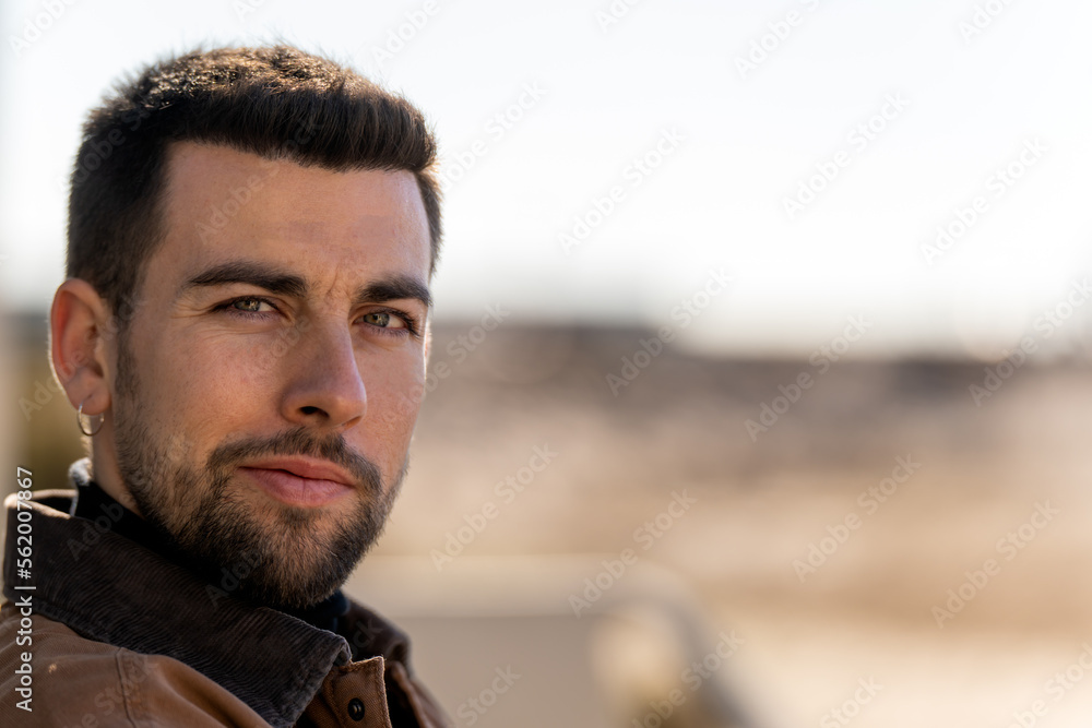 Portrait of positive bearded male with dark hair and earring in warm clothes looking at camera while sitting against blurred background