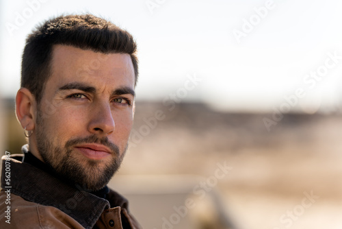 Portrait of positive bearded male with dark hair and earring in warm clothes looking at camera while sitting against blurred background
