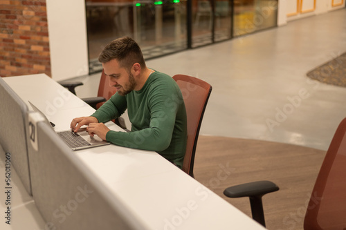 Caucasian bearded man working on laptop in modern coworking space. 