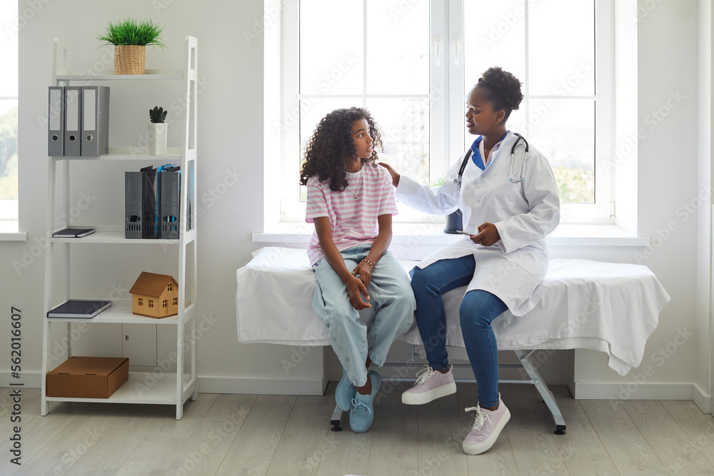 Caring, understanding doctor touches shoulder of excited teenage girl who is worried before medical examination. African American girl is sitting on examination couch and talking to female doctor. - obrazy, fototapety, plakaty 