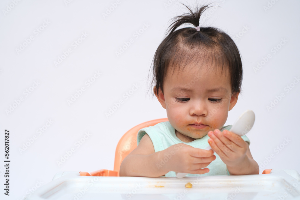 Cheerful baby child sitting and eats food by herself with spoon. Portrait of happy kid boy or girl in high chair. Smiling feeding delicious food concept. Isolate on white background. Cheerful baby child sitting and eats food by herself with spoon. Portrait of happy kid boy or girl in high chair. Smiling feeding delicious food concept. Isolate on white background.