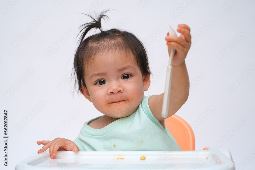 Cheerful baby child sitting and eats food by herself with spoon. Portrait of happy kid boy or girl in high chair. Smiling feeding delicious food concept. Isolate on white background. Cheerful baby child sitting and eats food by herself with spoon. Portrait of happy kid boy or girl in high chair. Smiling feeding delicious food concept. Isolate on white background.