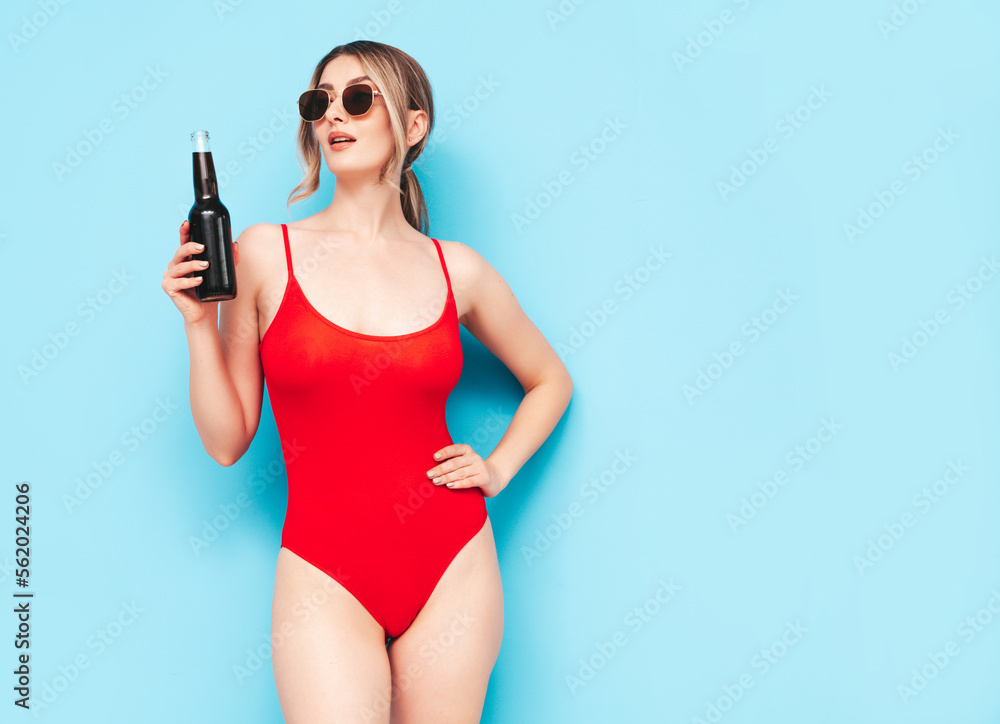 Young blond model in summer red swimwear bathing suit. Sexy carefree woman having fun and going crazy. Female posing near blue wall in studio. Drinking fresh cocktail smoozy drink from glass bottle
