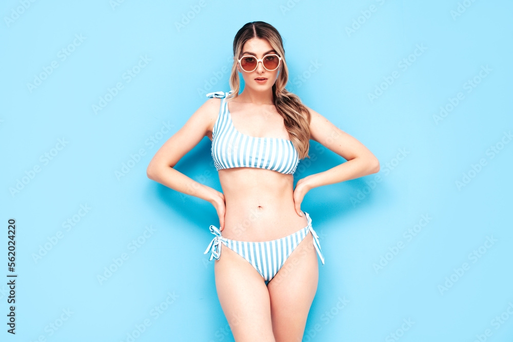 Obraz premium Young blond model in summer striped swimwear bathing suit. Sexy carefree woman having fun and going crazy. Female posing near blue wall in studio. Cheerful and happy. Perfect body. In sunglasses