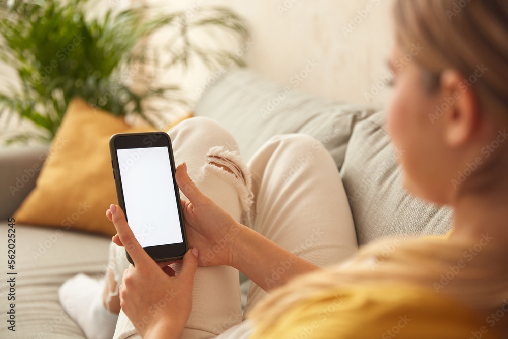 Woman Using Smartphone At Home. Blonde Girl Sitting On Sofa And Messaging In Cozy Living Room at Home. Female Person Doing Online Shopping, Browsing Internet and Checking Social Media 