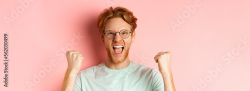 Close up of satisfied lucky redhead man winning, shouting from joy and making fist pump, celebratig victory, standing like champion over pink background photo