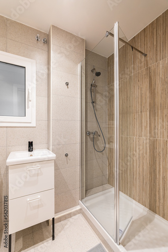Corner of bathroom with small shower area enclosed by transparent glass wall. Washbasin on table with drawers for bathroom accessories. Window in wall with frosted glass and white frame.