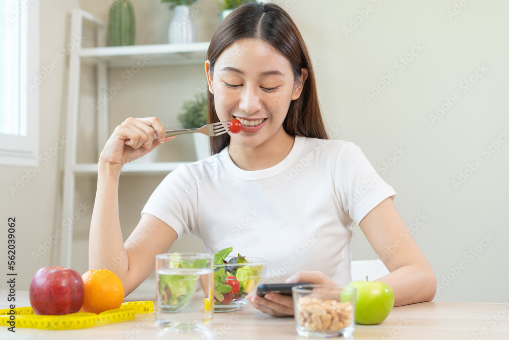 Dieting, smile asian young woman eating, holding fork at tomato, diet plan nutrition with fresh vegetables salad, enjoy meal while using smartphone. Nutritionist of healthy, nutrition of weight loss. Dieting, smile asian young woman eating, holding fork at tomato, diet plan nutrition with fresh vegetables salad, enjoy meal while using smartphone. Nutritionist of healthy, nutrition of weight loss.