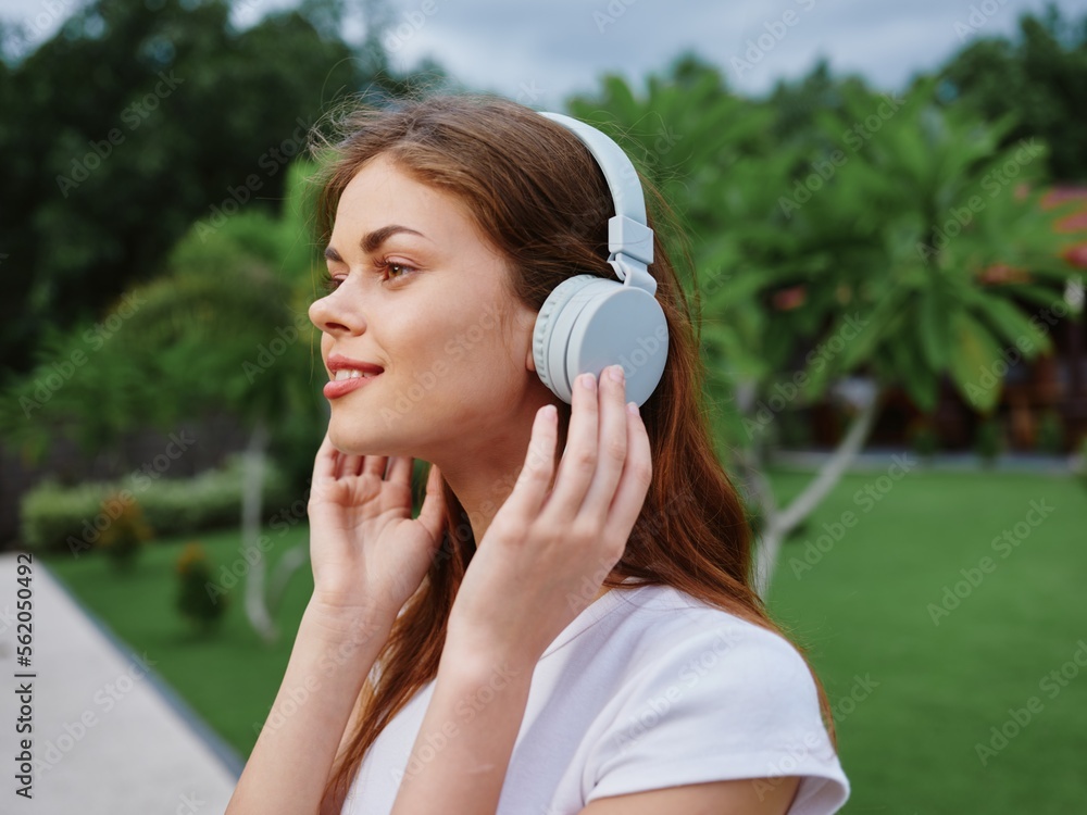 Woman portrait in headphones happiness smile in a white T-shirt listening to music and walking down the street, in front of palm trees Woman portrait in headphones happiness smile in a white T-shirt listening to music and walking down the street, in front of palm trees