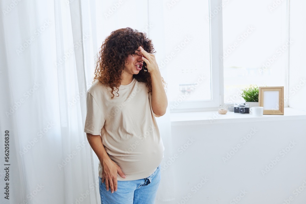 A pregnant woman stands in the house at the window, tired and has a headache, tears, crying from fatigue in a home T-shirt, the complexity of motherhood A pregnant woman stands in the house at the window, tired and has a headache, tears, crying from fatigue in a home T-shirt, the complexity of motherhood