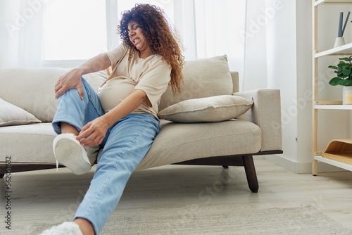 Pregnant woman sitting on the couch at home and wearing comfortable shoes for pregnancy with a problem due to the stomach, headache, pregnancy and motherhood difficulties, severe fatigue