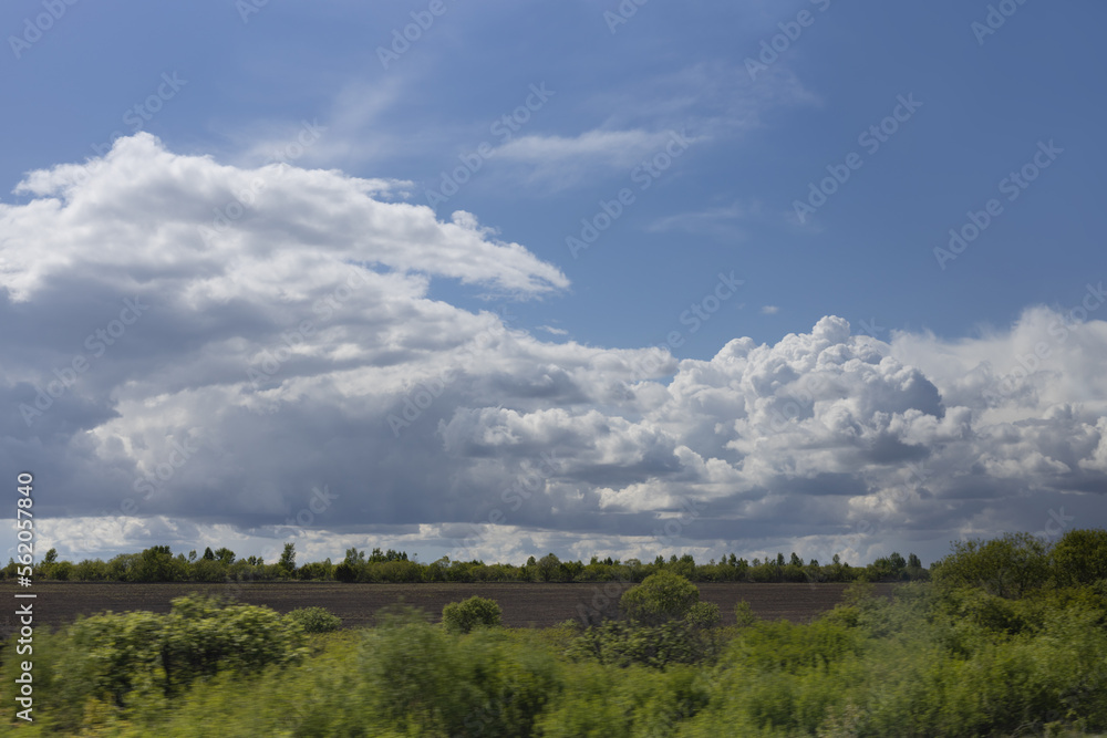 Bright early spring landscape with fluffy clouds in blue sky with sunbeams abow black arable land and lush green bushes. Beautiful outdoors nature.