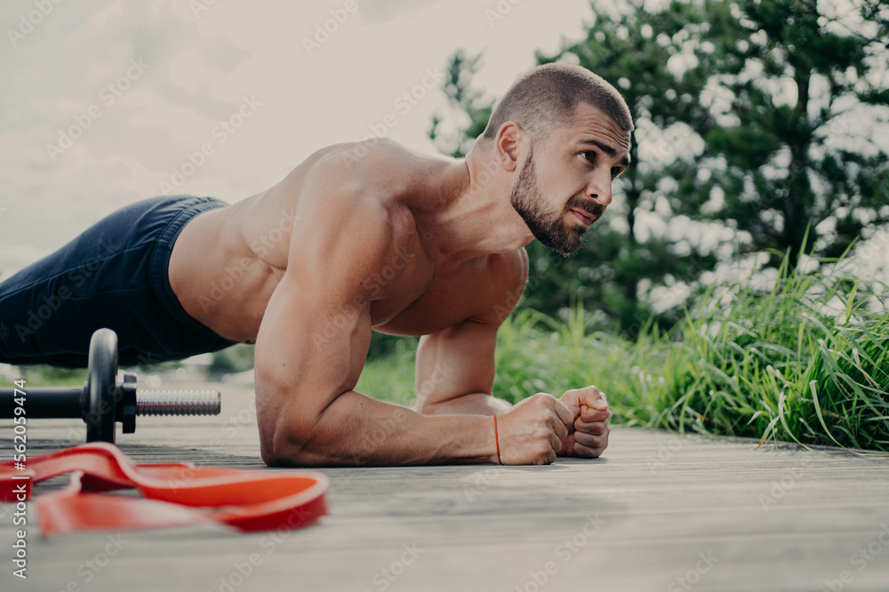 Obraz premium Photo of motivated bodybuilder does plank exercise, stands on elbows, uses sport equipment for workout, concentrated into distance, has muscular body, enjoys training outdoor. Morning fitness concept