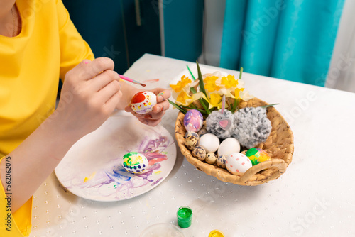 Top view of female hands in close-up, decorating eggs for Easter. On the table there is a wicker basket with a toy rabbit and a daffodils. Concept of traditional celebrations and creativity