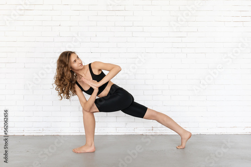 Young woman practicing yoga, doing parivritta parshvakonasana exercise, side angle unfolded pose, exercising in black sportswear near white brick wall