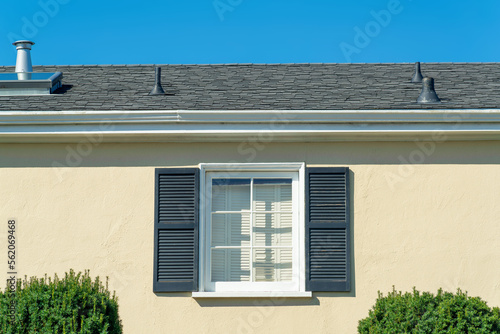 Lone window on beige house facade with green shutters and white accent color with dark roof tiles and vents
