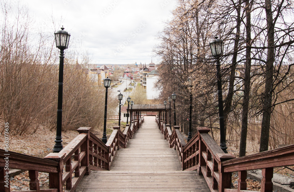 Obraz premium Wooden high staircase for descent from the mountain to the city. Lanterns, massive railings. Old trees and shrubs. Below you can see the streets and houses of the townspeople. Early spring.