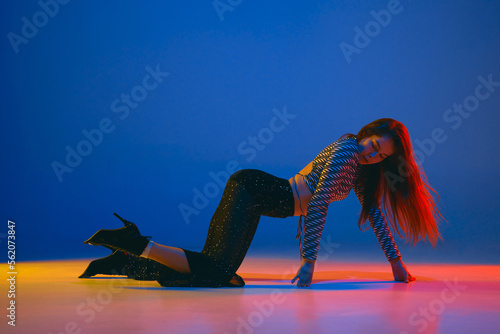 Self-expression. Young girl dancing high heel dance in stylish clothes over blue background in neon light. Concept of modern dance style