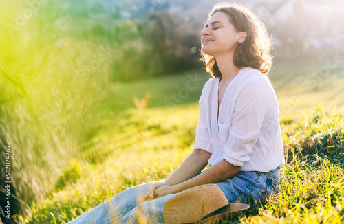 Obraz na płótnie Happy pretty young cheerful woman in white shirt sitting on grass enjoying sunny spring day with eyes closed w salonie