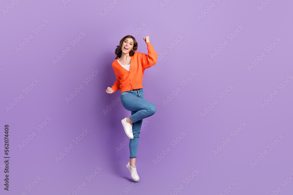 Full length photo of lucky cheerful lady wear orange cardigan jumping high rising fists empty space isolated violet color background