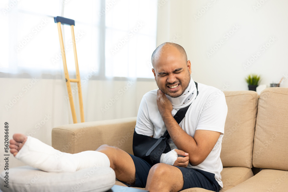 Man suffered pain from accident fracture broken bone injury with leg splints in cast neck splints collar arm splints sling support arm in living room. Social security and health insurance concept.