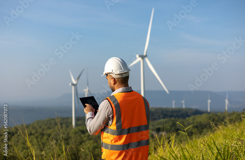 Engineer India man working with tablet at windmill farm Generating electricity clean energy. Wind turbine farm generator by alternative green energy. Asian engineer checking control electric power
