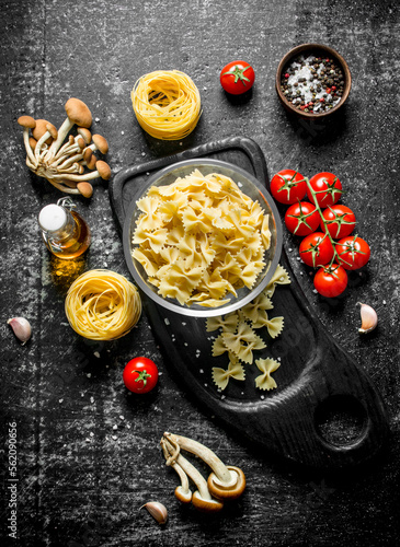 Raw farfalle and tagliatelle paste in a bowl on a cutting Board with mushrooms, tomatoes and spices.