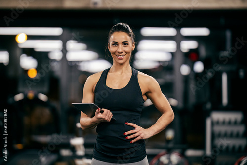 Fototapet A fit muscular female personal trainer is holding tablet in her hands and smiling at the camera in a gym